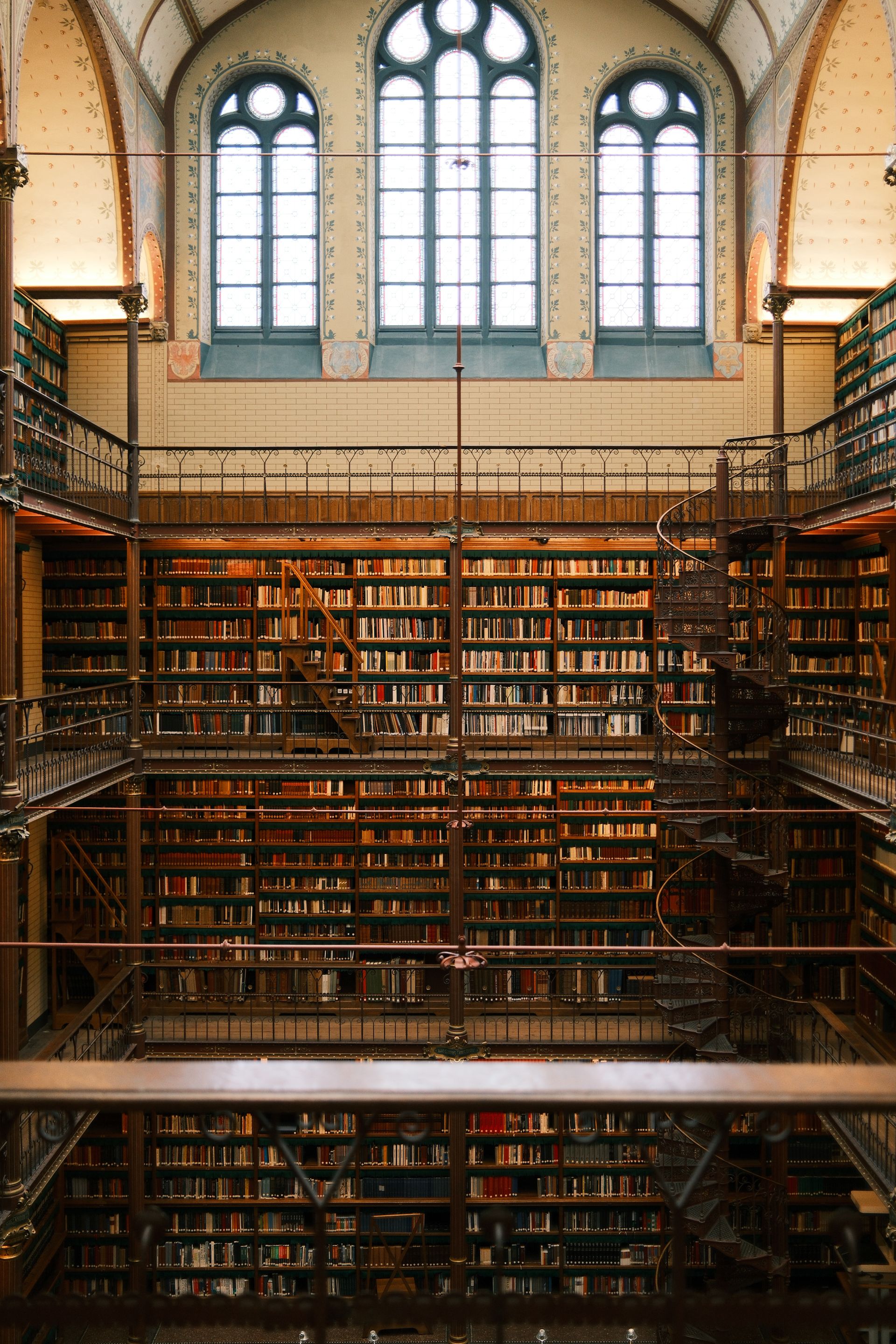 A grand multi-level historic library with arched windows, a spiral staircase and towering bookshelves